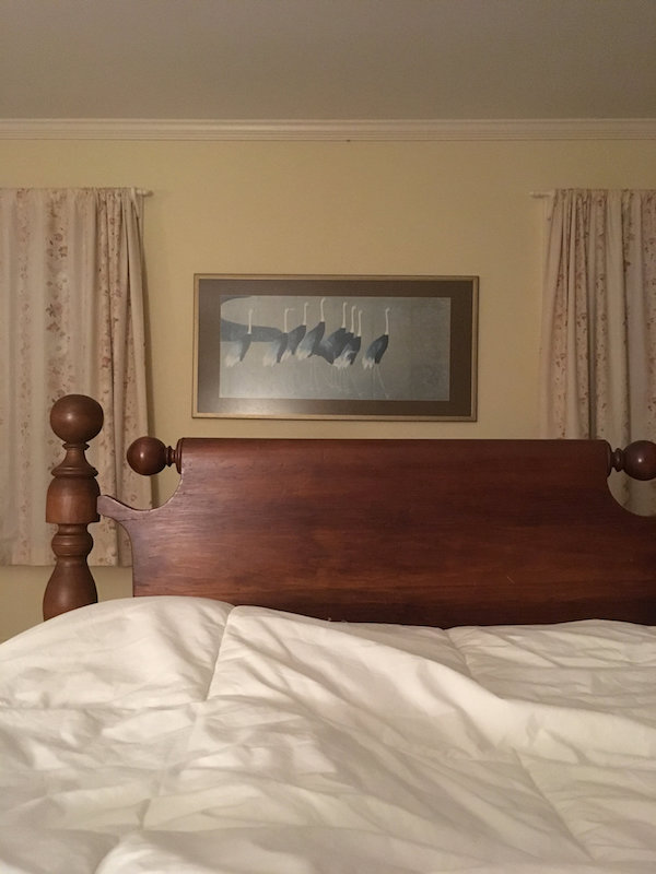 showing the foot of a wooden bed with a crane photo on the wall behind it.