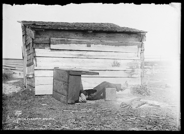 black and white image of a person asleep on their back in front of a little midwestern looking cabin