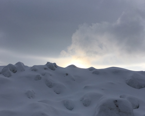 photo of a wallk of snow with the sun peeking out from behind it with some wispy clouds being lit by the sun.