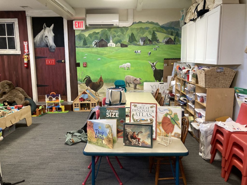 View of the children's room in the library which features a table highlighting books about dinosaurs and has a rural mural on the wall including a field with cows and also a horse stall with a lifesize illustration of a horse in it.